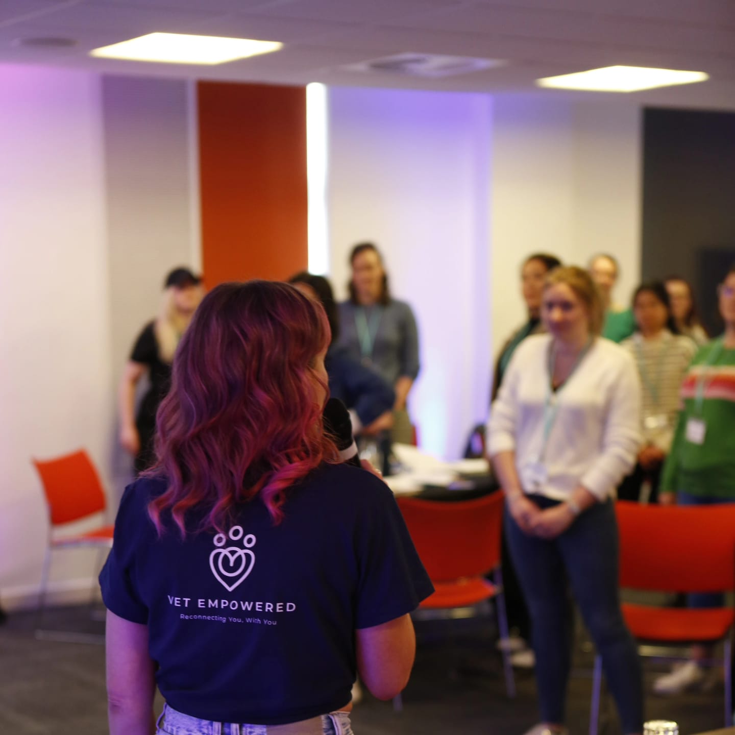 A Vet Empowered facilitator in a branded t-shirt, seen from behind, speaking with a seated group of veterinary professionals